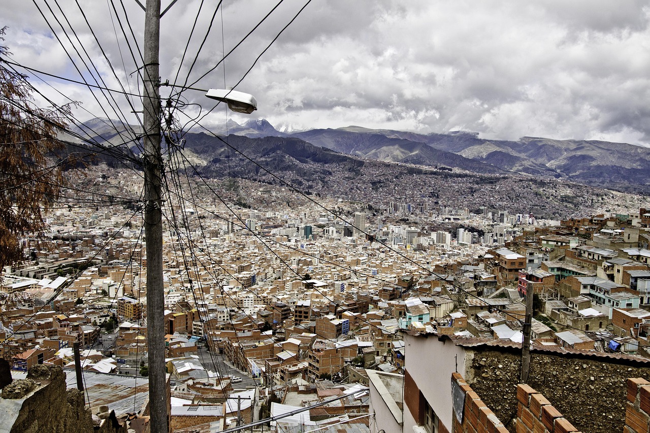 A wide panoramic view of the city of La Paz, Bolivia, with dense buildings in a valley surrounded by high mountains under a cloudy sky.