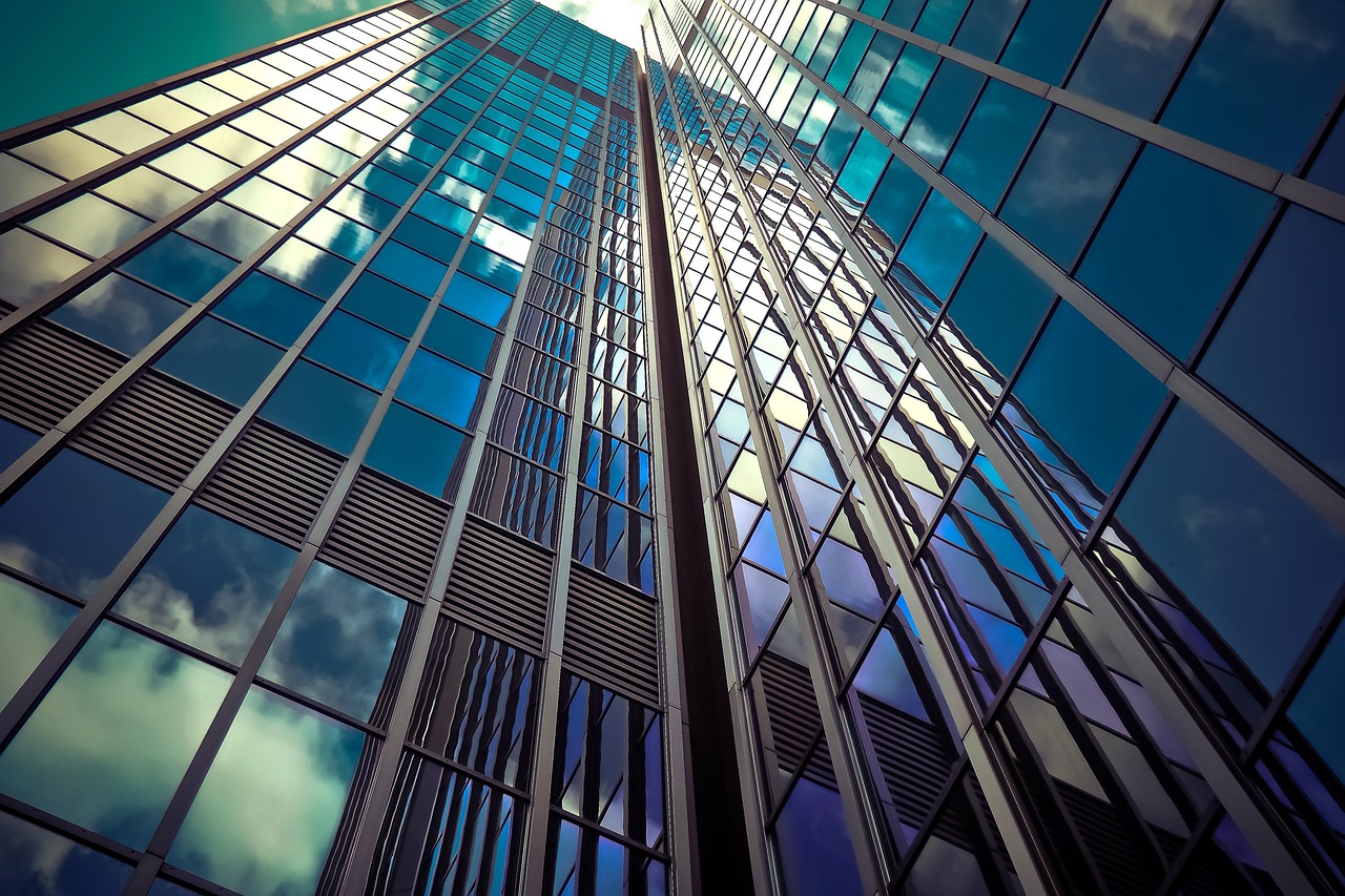 A low-angle view of two modern glass skyscrapers reaching toward a bright sky with light clouds reflected in the windows.