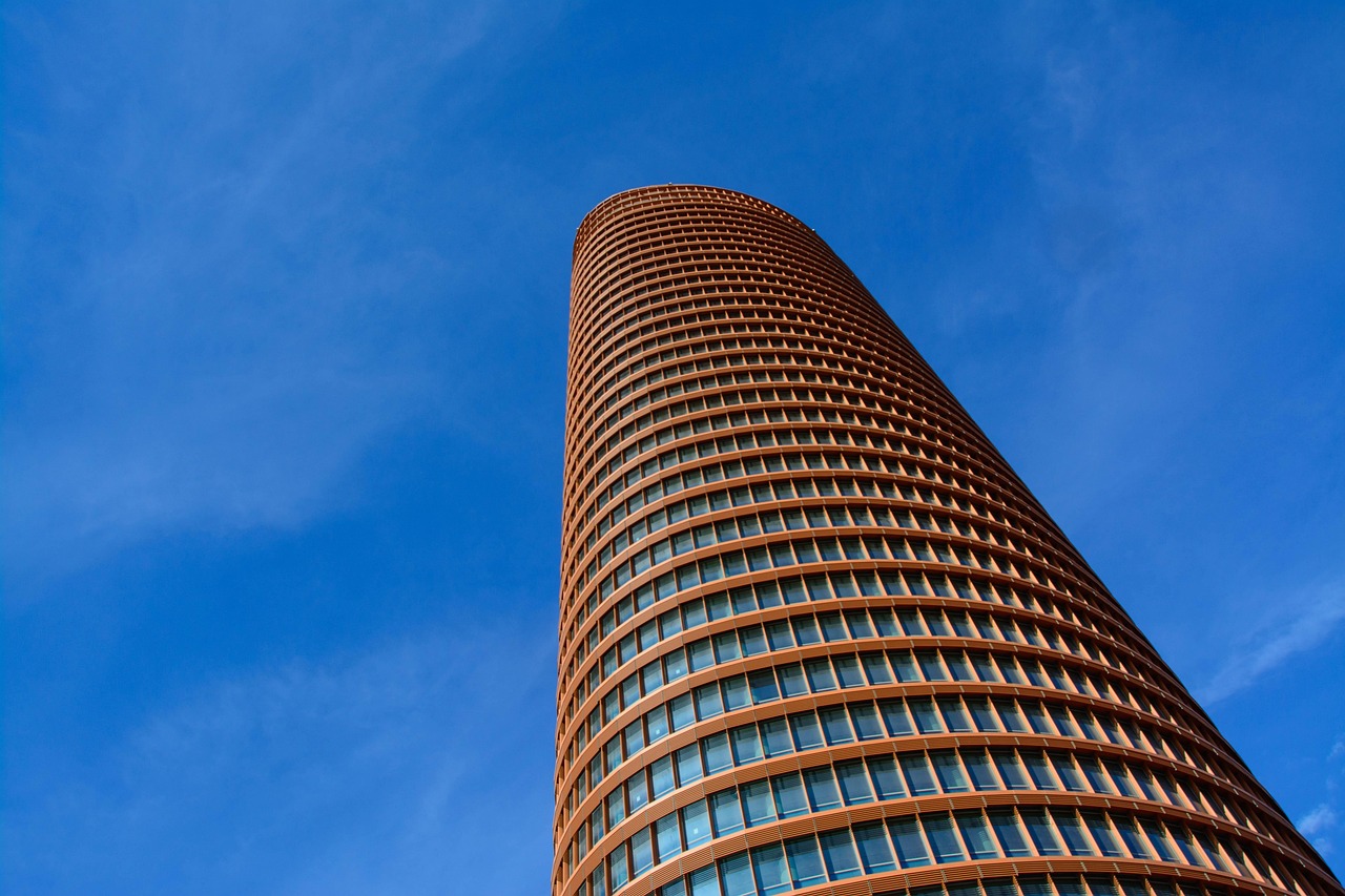 A low-angle view of a tall, modern cylindrical office building with many windows under a clear blue sky.