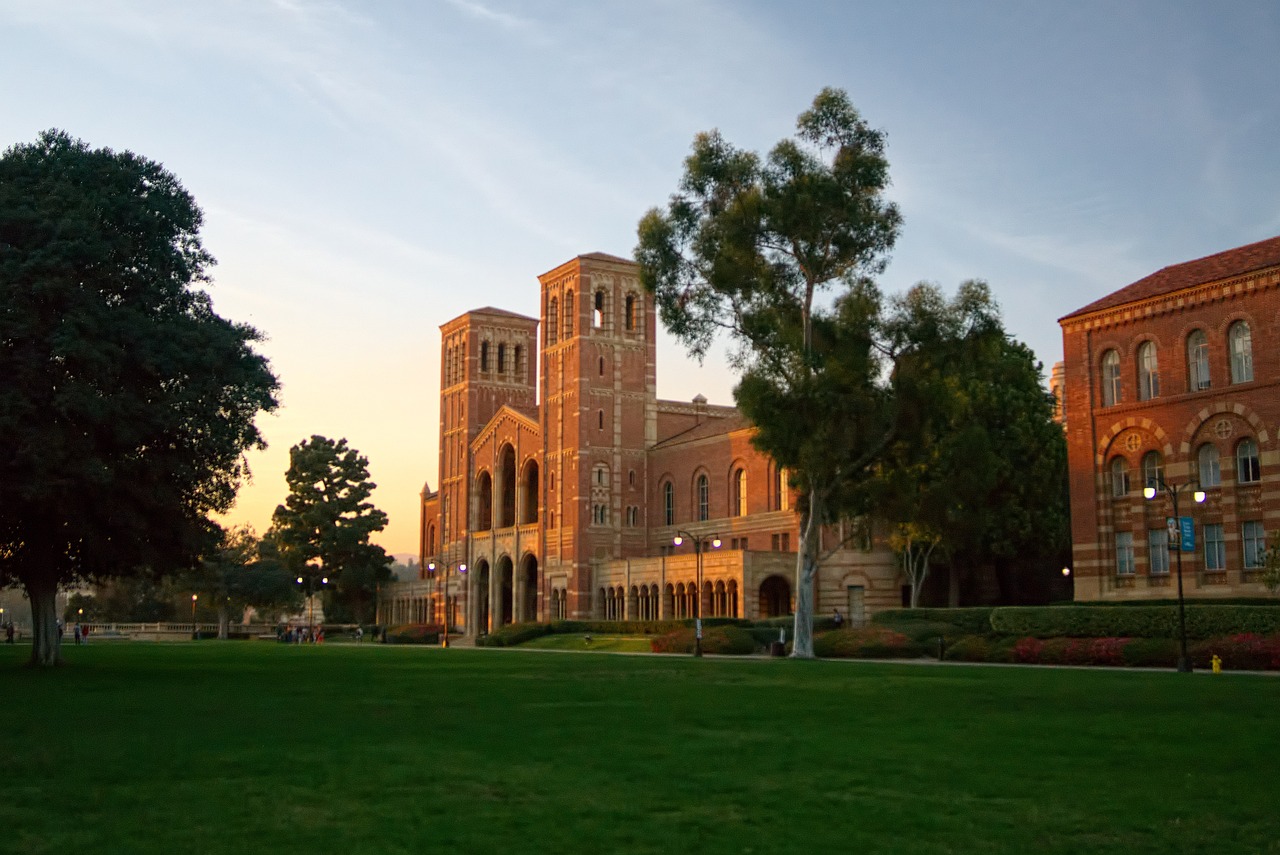 A large university campus with a grand brick building featuring two towers, set behind a lush green lawn during a clear sunset.