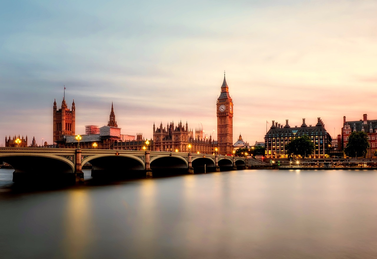 A wide landscape view of the Palace of Westminster and Big Ben in London at sunset, reflected in the River Thames.